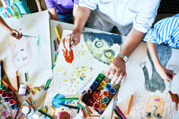 top view of childrens drawings on table in art class with group of people painting and teachers hand