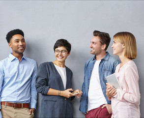 Four mixed race people or creative workers meet together, have intriguing discussion, share their ideas on improvement business project, use modern cell phones and tablet, isolated over grey wall