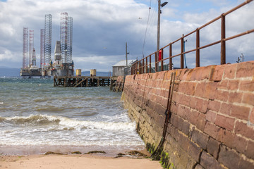 Waves along a stone pied with a rusted ladder and oil platform in the background