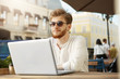 © Cookie Studio - Young mature red-haired bearded guy in stylish clothes and tan glasses sitting outside in sunny summer day, working on team project via laptop and feeling happy.