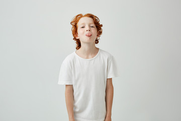 Wall Mural - Close up portrait of beautiful ginger kid in white t-shirt showing tongue in camera posing for family photo. Lifestyle concept.