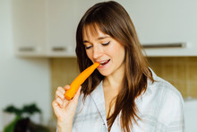 Pretty Young Woman In Pajamas Eating Carrot In The Kitchen