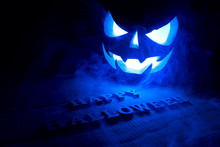 The Concept Of Halloween. Glowing With Icy Blue Light Angry Terrible Pumpkin. Jack Lantern In The Dark, On A Wooden Background With A Voluminous Inscription Of Happy Halloween With Smoke
