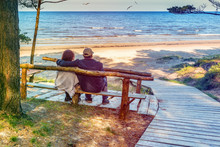 Happy Elderly Couple Resting On The Wooden Bench And  Looking At The Distance Of The Baltic Sea. Image Slightly Toned For Inspiration Of Retro Style.