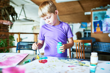 Wall Mural - Portrait of blonde little boy painting pictures  carefully, standing at table in art studio