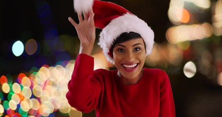 Wall Mural - Cheerful young woman modeling Santa Claus hat, smiling and laughing. Portrait of cute black female playing with festive hat in outdoor setting with pretty bokeh lights