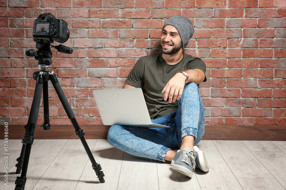 Young blogger with laptop recording video against brick wall background