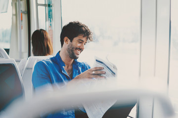 Young model look male is reading a newspaper while sitting in a city tramway beside a window. Bearded man with dark curly hair wearing light blue shirt is going to the office by a public transport.