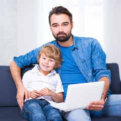 Wall Mural - computer and internet concept - young father and little son sitting on sofa with laptop