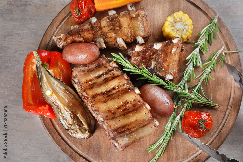 Wooden board with grilled ribs and vegetables on table