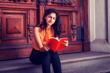 Way to Success. Power of Reading. East Indian American college student studying in New York, sitting on steps in front of vintage style library door way, looking down, reading red book, thinking..