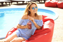 Beautiful Young Woman With Fresh Smoothie Sitting On Bean Bag Chair Near Swimming Pool