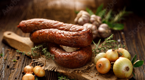 Fototapeta na szkło Wędzona kiełbasa na drewnianym rustykalnym stole z dodatkiem świeżych aromatycznych ziół i przypraw, naturalny produkt z gospodarstwa ekologicznego, wytwarzany tradycyjnymi metodami