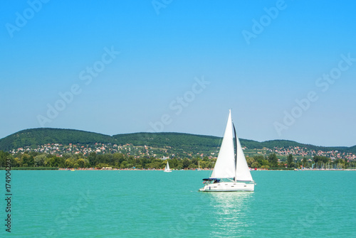 A view from a ship to bright Balaton lake water and a white yacht with a town, forest and mountains at the background on sunny summer day, Hungary Obraz na płótnie
