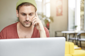 Wall Mural - Always in touch. Portrait of hipster guy with stubble surfing internet on generic portable computer and talking on mobile phone, having concentrated serious expression, working at coffee shop