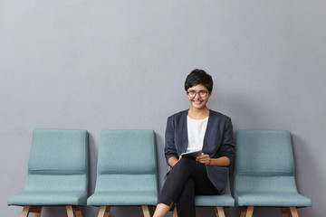 Indoor shot of positive business lady comes in polyclinic, makes appointemnt to doctor, sits in queue, waits for turn, communicates with partners online as uses modern tablet. Technology, job