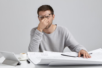 Stess, overwork and deadline. Tired unshaven young architect freelancer massaging nose bridge , having headache while working in big architectural project at home office, feeling exhausted and sleepy