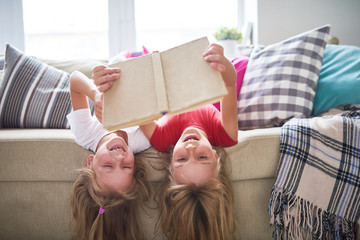 Portrait of two happy blonde sisters having fun at home reading book together lying upside down on sofa and smiling