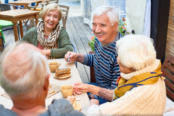 Joyful elderly friends having tea party at lovely outdoor cafe: they enjoying pastry and fragrant coffee and chatting animatedly with each other