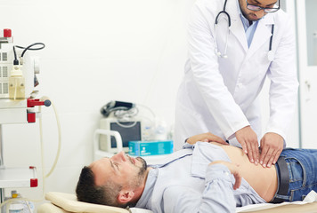 Wall Mural - Concentrated young physician doing abdominal palpation of middle-aged patient suffering from pain, interior of modern office on background