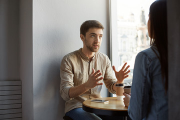 Serious unshaved dark haired man sitting in cafeteria with client, talking and gesticulating with hands, trying to make clear some details of commission he received.