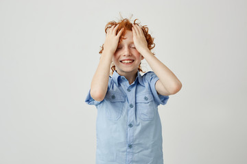 Wall Mural - Cute little boy with red wavy hair and closed eyes holding hands on forehead, laughing and being shy about saying stupid joke to friend.