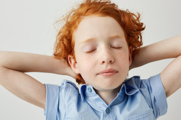 Wall Mural - Close up of beautiful sleepy ginger boy with curly hair and freckles holding hands behind head with closed eyes being tired after school classes.