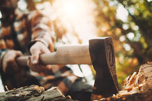 Strong Lumberjack In Plaid Shirt Holding Ax In His Hands And Chopping Tree In Forest. Axe Close Up. Blurred Background, Lens Flare Effect