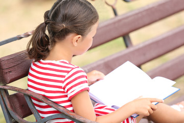 Wall Mural - Cute little girl reading book while sitting on bench in park