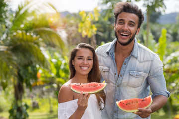 Poster - Portrait Of Happy Couple Eating Watermelon Together Cheerful Diverse Man And Woman Holding Slice Of Watermelon Outdoors Over Palm Trees Background