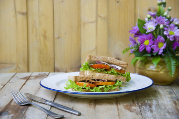 Two fresh pieces of ham sandwiches with bacon, lettuce and tomato in a dish on a wooden table.