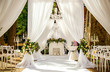 Place for wedding ceremony in white color ,with white fireplace and chandeliers decorated with flowers and white cloth and wooden chairs for guests on each side outdoors.