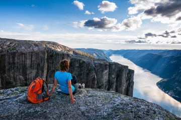 Wall Mural - A woman is sitting on the edge of cliff on the way to boulder (Kjeragbolten) stuck in between the mountain crevices of Kjerag above a fjord, near Lysebotn, Norway. The feeling of complete freedom