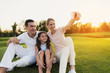 © VadimGuzhva - Family doing selfie on gray smartphone sitting on grass on golf course on sunset background
