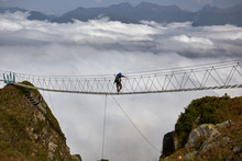 Man Walking On Suspension Bridge And Looking At Cloudy Mountains Below.