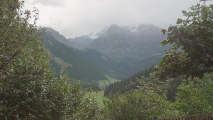 Wall Mural - Handheld 4k shot of green alpine valley and small village in the Switzerland. Stunning fields on a cloudy day and famous stunning touristic town with high cliffs in background.