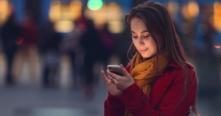 Young Woman Using Smartphone at Dusk in the City. 4K SLOW MOTION. Millennial Girl reading on cell phone, using app, communicating in social media, texting. Blurred Night Street lights background.