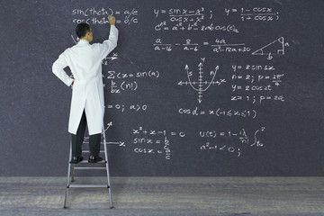 Scientist of genius standing on a ladder writing complex mathematical formulas with white chalk on large blackboard.