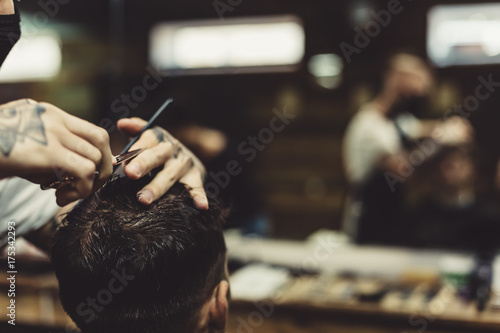 Fotografie, Obraz Anonymous stylish barber with tattoos cutting hair of male client in chair