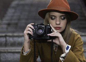 Portrait of young woman dressed in hat and coat looking down to photo camera on stairs background