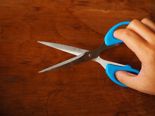 Right hand holding, opening blue plastic with stainless steel blade scissors, with red brown wooden texture table background