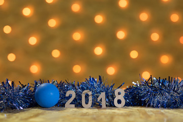 Wall Mural - Blue tinsel and a ball with figures of 2018 on a table on the background of a New Year's garland with golden lights