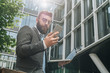 © foxyburrow - Young bearded businessman stands against modern building, uses smartphone and holds a laptop in his hand. Man works remotely. Distance job. Online marketing, education,training,e-commerce,e-learning.