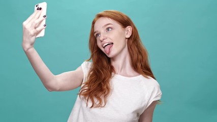 Sticker - Happy ginger woman in t-shirt making selfie on smartphone and showing many funny gestures at camera over turquoise background