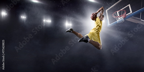 Plakat Gracz koszykówki robi slam dunk na dużej profesjonalnej arenie. Gracz leci w powietrzu z piłką. Gracz nosi ubrania niemarkowe.
