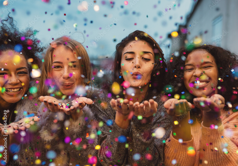 Young women blowing confetti from hands. Friends celebrating outdoors in evening at a terrace. - obrazy, fototapety, plakaty 