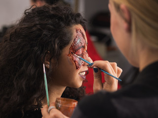 Make-up artist make the girl halloween make up in studio.Halloween face art.Woman applies on professional greasepaint on the face of girl.War-paint with blood, scars and wounds.