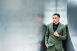 © Alexander Image - Portrait of young  American Businessman in New York, wearing green suit, black undershirt, hand on front, standing by sliver metal wall, looking away, thinking. Color filtered with dart blue tint..