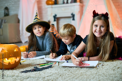 Zdjęcie XXL Grupowy portret rozochoceni dzieci jest ubranym Halloweenowych kostiumy zbierał wpólnie przy wygodnym pokojem dekorującym dla Halloween i rysuje kolorowych obrazki, dziewczyny patrzeje kamerę z szerokimi uśmiechami
