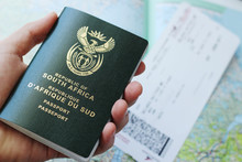 A South African Passport In A Man's Hand With A Plane Ticket And Map In The Background. This Image Can Be Used To Represent Emigration Or Travelling. This Image Has Selective Focusing. 